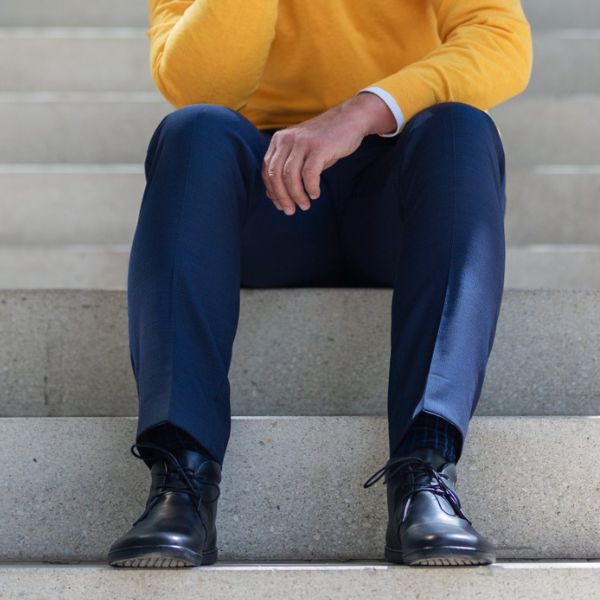 Eine Person in einem leuchtend gelben Pullover und dunkelblauen Hosen sitzt auf Betonstufen. Schwarze Lederschuhe sind ordentlich an ihren Füßen geschnürt. Die Person stützt ihre Unterarme auf ihre Oberschenkel und hat die Hände locker gefaltet. Die Betonstufen bilden einen neutralen grauen Hintergrund.