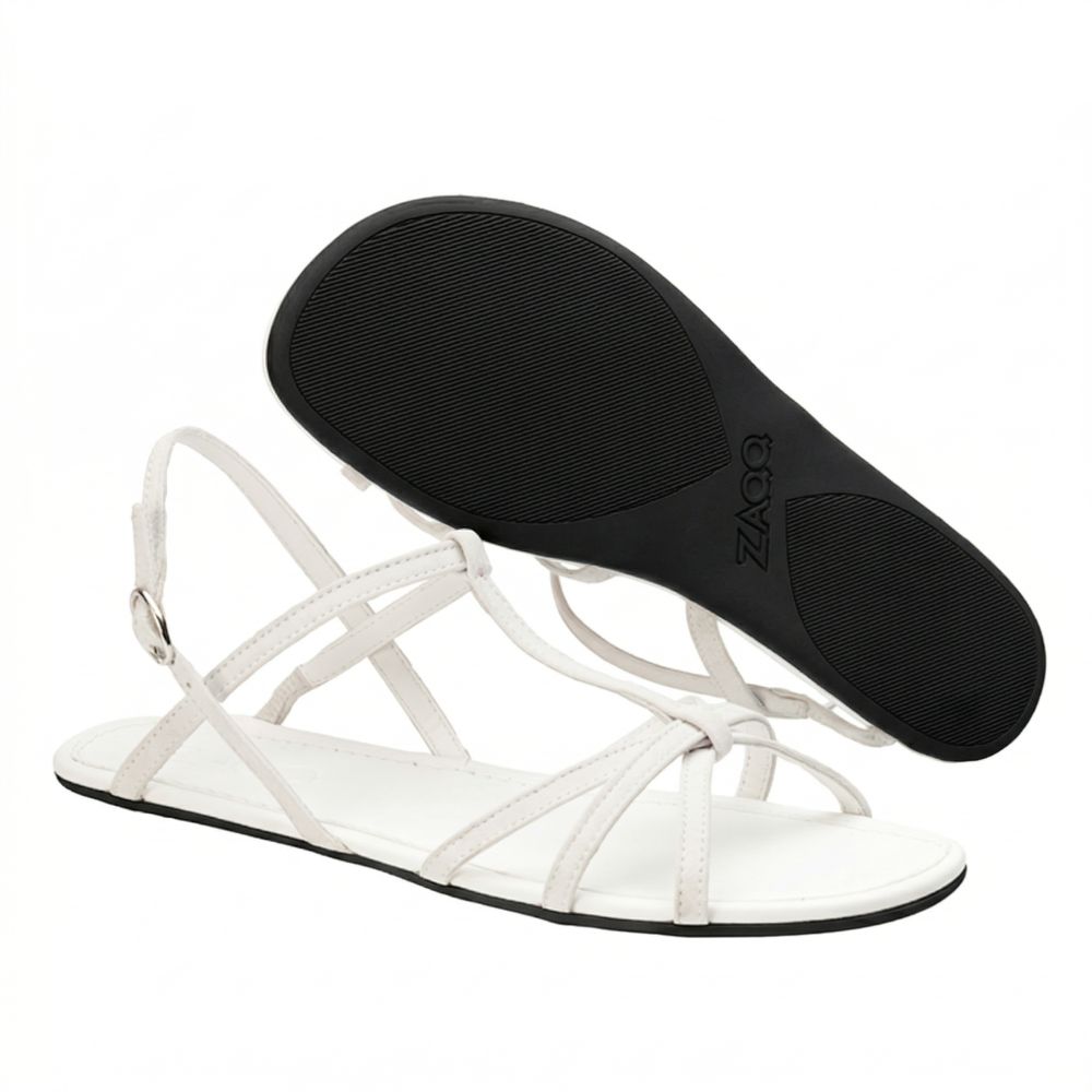 Ein Paar weiße Riemchensandalen ZAQQ QADA aus hochwertigem Nappaleder mit flacher Sohle. Eine Sandale steht aufrecht und zeigt ihre elegante Schnalle, während die andere umgedreht die strukturierte Sohle freigibt – die perfekte Mischung aus Stil und Komfort.