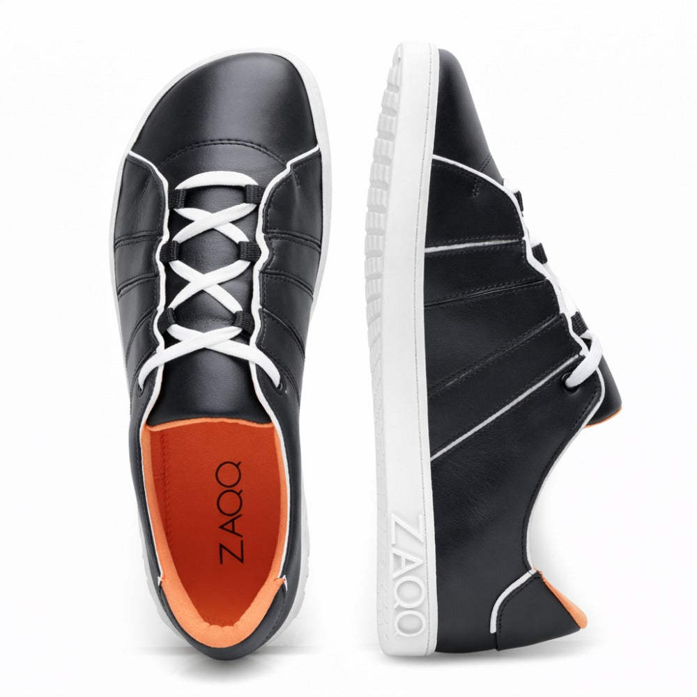 Die QQQ Low Black White Sneaker von ZAQQ zeichnen sich durch ein elegantes schwarzes Lederdesign mit weißen Sohlen und Schnürsenkeln aus. Ein Schuh ist von oben zu sehen und zeigt seine rote Innensohle und das markante ZAQQ-Branding, während der andere von der Seite zu sehen ist und sein stilvolles Design und seinen Komfort hervorhebt.